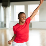 A young Black adult wearing athletic clothes in a dance studio reaches one arm up in an active stretch